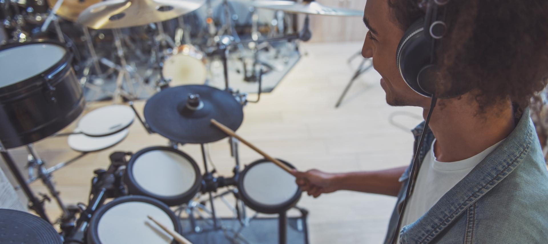 A person playing an electronic drum kit in a studio setting, wearing headphones and holding drumsticks.