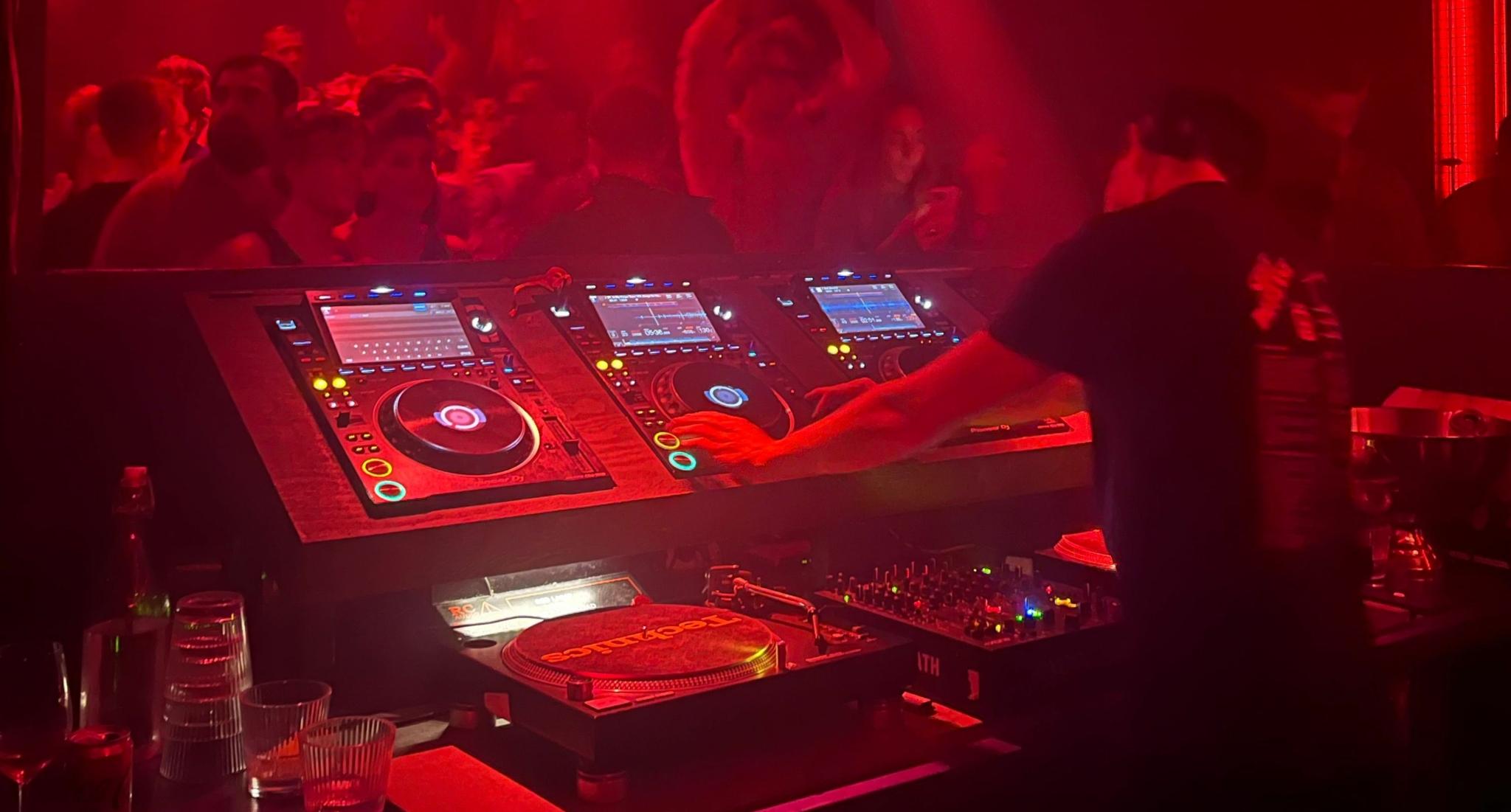 Red-lit nightclub DJ booth with DJ decks, mixer, and crowd