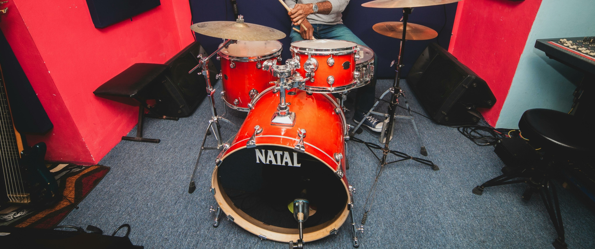 A drummer playing a bright orange Natal acoustic drum kit in a colorful studio setting.