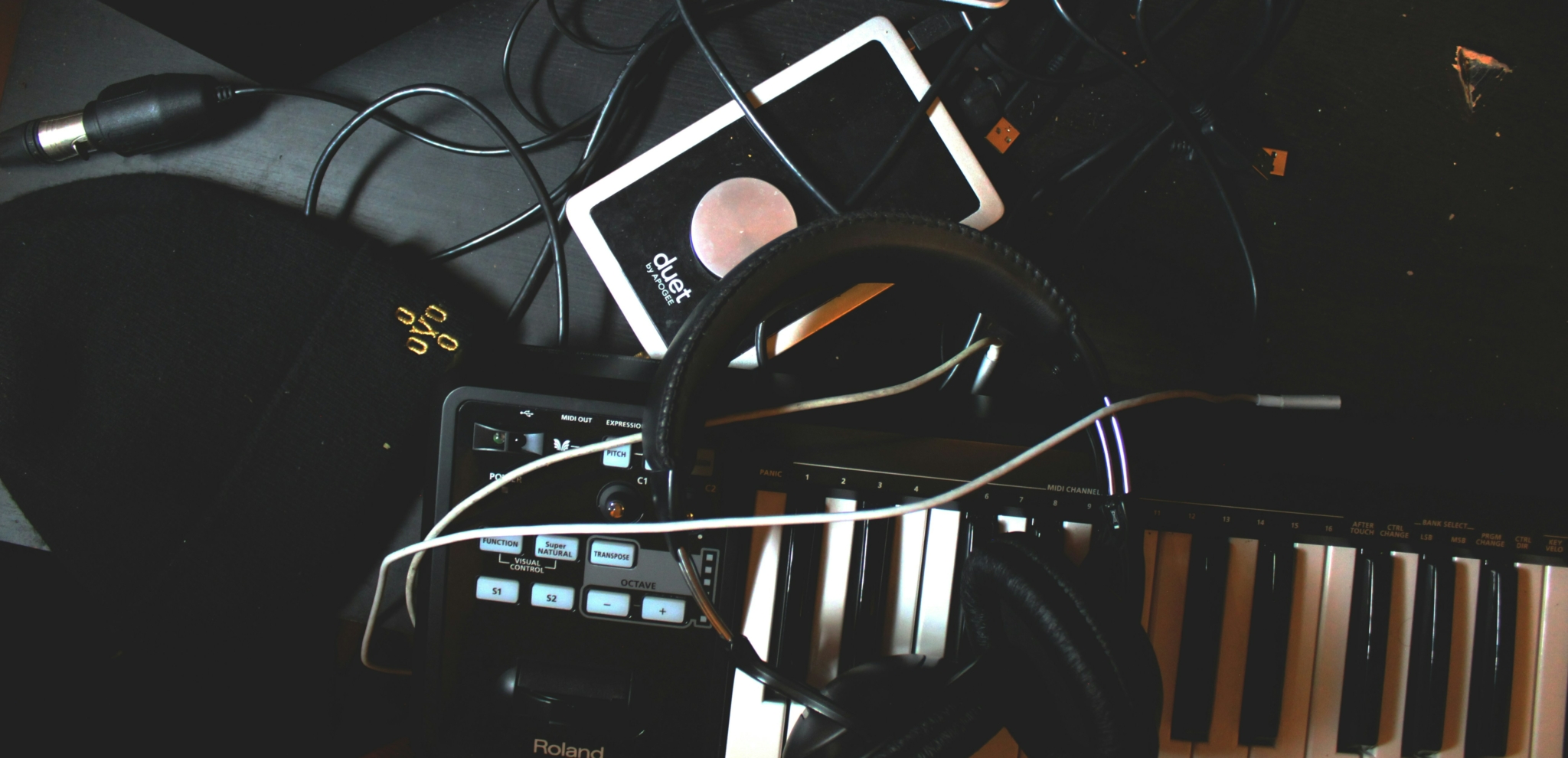 A close-up of music production equipment, showcasing a Duet audio interface, Roland MIDI keyboard, and headphones amidst tangled wires.