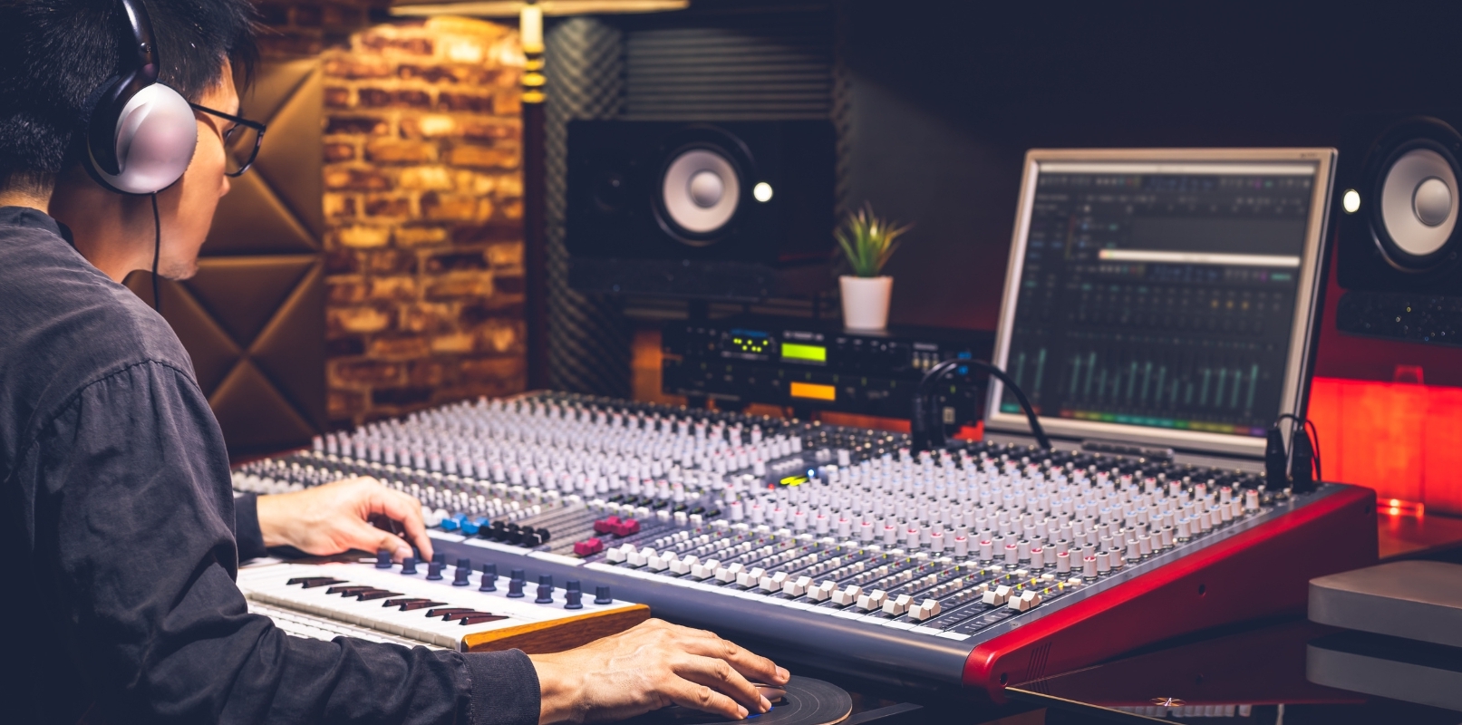 Music producer mixing on a large console with studio monitors and computer display in the background.
